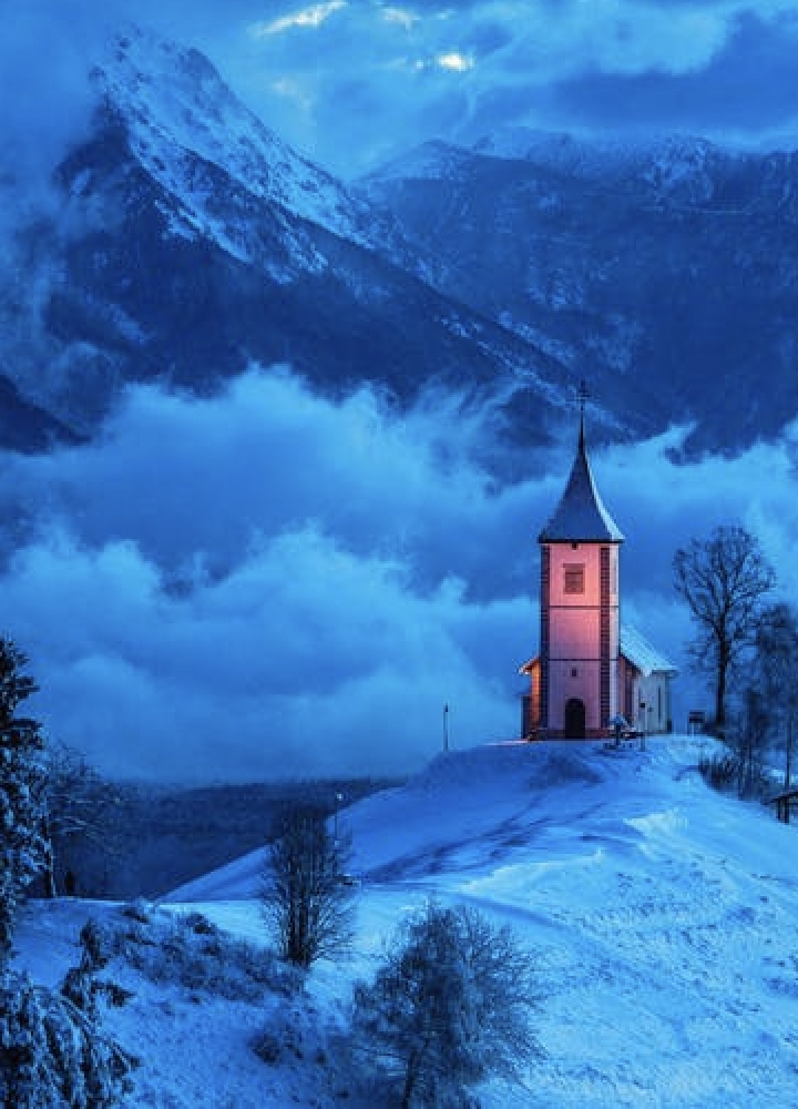 A small church on top of a tall hill surrounded by clouds and snow caped mountains.